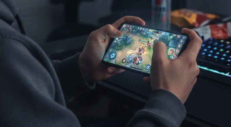 Seorang sedang bermain Mobile Legends di ponselnya dengan latar belakang monitor komputer dan camilan